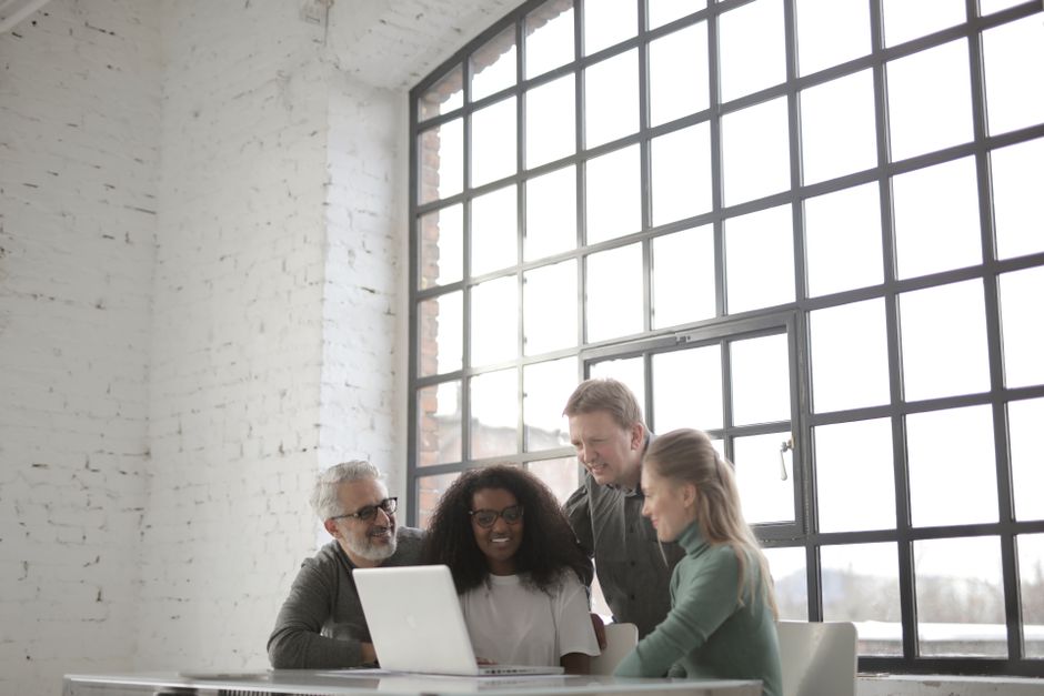 AI startup news - Positive focused multiracial coworkers gathering together near table with laptop in workplace at industrial building during remote work on team against big window at daytime